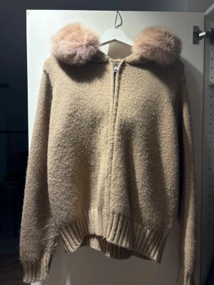 Alessa jacka - Mysig beige stickad hoodie med dragkedja framtill och stor huva kantad med fluffig  päls. Tröjan har ribbade muddar vid ärmslut och nederkant, perfekt för kalla dagar. Materialet känns mjukt och värmande.