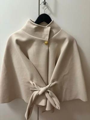 Beige poncho med knyt och guldknapp - Stilren beige Poncho med hög krage, dekorativ guldknapp vid halsen och knytband i midjan. Jackan har en elegant, vid siluett och är tillverkad i ett mjukt tyg som ger en exklusiv känsla. Perfekt för dig som vill ha en chic och enkel look. Helt ny och oanvänd från missäy.