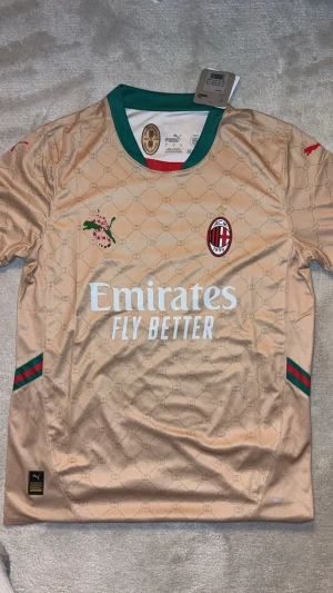 Beige AC Milan x Puma matchtröja - Snygg beige AC Milan matchtröja från Puma med gröna och röda detaljer, samt klubbmärke och sponsortryck framtill. Tröjan har korta ärmar, rund halsringning och diskret mönster i tyget. Perfekt för fotboll eller träning.