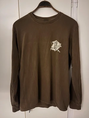 Brun långärmad tröja från Stussy - Brun långärmad tröja från Stussy med tryck på bröstet och stort grafiskt tryck på ryggen där det står 'Stussy International'. Skön passform och rund halsringning, perfekt till streetwear-stilen.