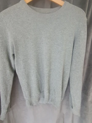 Ljusgrå stickad tröja unisex - En stilren ljusgrå stickad tröja med rund halsringning och långa ärmar. Tröjan har ribbade muddar vid ärmslut och nederkant. Perfekt basplagg i mjukt material som passar till det mesta.