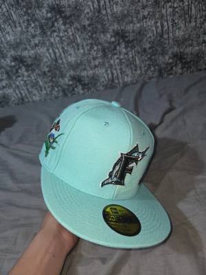 Turkos Marlins keps från New Era - Snygg turkos keps från New Era med Miami Marlins-logga framtill. Kepsen har broderade fjärilar och blommor på sidorna för en unik look. Flat skärm och klassisk 59FIFTY-modell. Perfekt för dig som vill sticka ut med din stil.