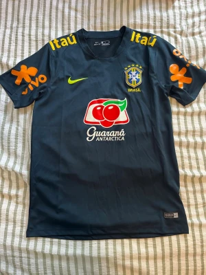 Brasil tröja - Snygg mörk grön träningströja från Nike i storlek S med Brasil-logga och flera färgglada sponsortryck i gult, orange, vitt och rött. Korta ärmar och rund hals. Tillverkad i lätt och ventilerande material, perfekt för fotbollsträning eller sport.