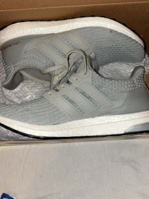 Grå Adidas Ultra Boost  - Snygga grå Adidas Ultra Boost sneakers med stickad ovandel och klassiska tre ränder på sidan. Skorna har en vit Boost-sula för extra komfort och svart yttersula. Perfekta för dig som gillar stilrena och bekväma sneakers.