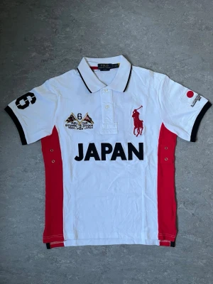 Polo Ralph Lauren Japan piké - Säljer en snygg vit pikétröja från Polo Ralph Lauren med röda sidopaneler och stora broderade detaljer. Tröjan har svart text 'JAPAN' framtill, klassisk krage med svarta kanter och knappar, samt broderad logga och patchar på bröstet och ärmen.