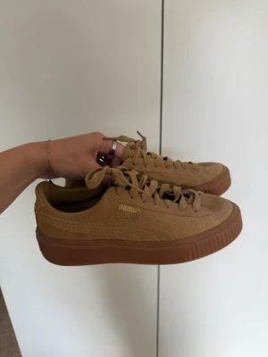 Beige sneakers från Puma i mocka - Snygga beige sneakers från Puma med ovandel i mocka och tjock gummisula i brunt. Klassisk låg modell med snörning och diskret guldfärgad Puma-logga på sidan. Perfekta för en clean och stilren look.