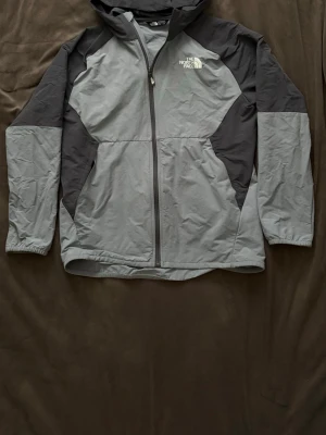Grå vindjacka från The North Face - Snygg grå vindjacka från The North Face med svarta detaljer på axlar och sidor. Jackan har huva, hel dragkedja och två sidofickor. Materialet är lätt och vindtätt, perfekt för att skydda mot blåst. Logotypen är tryckt på bröstet och ryggen.