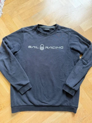 Svart Sail Racing sweatshirt - Svart sweatshirt från Sail Racing med tryckt logga i vitt på bröstet. Klassisk rund halsringning och ribbade muddar vid ärmslut och nederkant. Perfekt för en sportig och avslappnad stil.