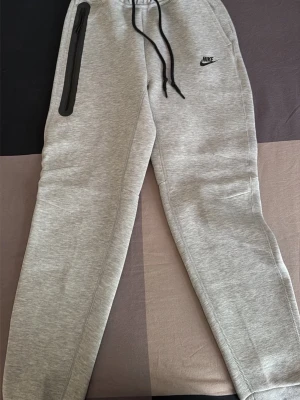 Grå mjukisbyxor från Nike - Snygga ljusgrå mjukisbyxor från Nike med svart logga på vänster sida. Byxorna har dragsko i midjan och en svart dragkedja på ena fickan. Perfekta för chill eller träning, riktigt sköna och stilrena.