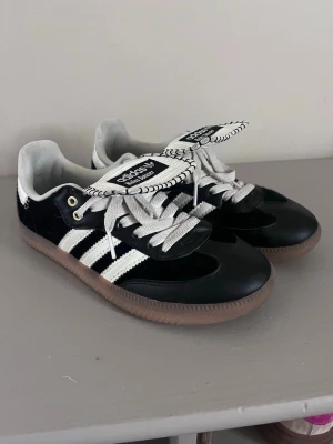 Adidas Wales Bonner svart/vit sneakers - Snygga Adidas Wales Bonner sneakers i svart och vitt med klassiska tre ränder på sidan. Skorna har en unik oversized plös med kontrastsömmar och Wales Bonner-logga. Ovandelen är i mocka och läder, och sulan är i gummi. Perfekta för dig som gillar streetwear och vill sticka ut.
