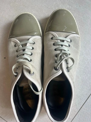 Grå sneakers från Lanvin - Snygga ekte grå sneakers från Lanvin med mocka på ovandelen och glansig tå i plast. Skorna har vita snören, svarta öljetter och en låg silhuett. Perfekta för dig som gillar stilrena och unika detaljer på dina sneakers.