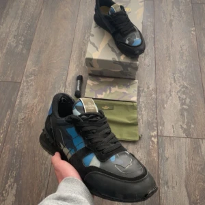 Valentino svart/blå camo sneakers - Snygga sneakers från Valentino med svart och blå camo-mönster, svarta skosnören och detaljer i mocka. Märkeslogga på plösen och cool design som sticker ut. Perfekt för dig som vill ha något unikt och trendigt.