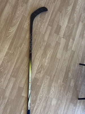 Bauer Vapor Hyperlite klubba - Säljer en Bauer Vapor Hyperlite hockeyklubba med svart och grått rutmönster på bladet och detaljer i vitt, gult och rött på skaftet. Klubban är lätt och tillverkad i kompositmaterial, perfekt för dig som vill ha snabb puckhantering.