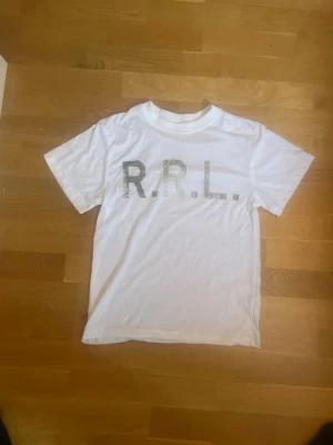 Vit t-shirt med RRL-tryck - Vit t-shirt från RRL med stort grått tryck på bröstet. Klassisk rund hals och korta ärmar. T-shirten är gjord i mjuk bomull och har en enkel, clean look som passar perfekt till jeans eller shorts.