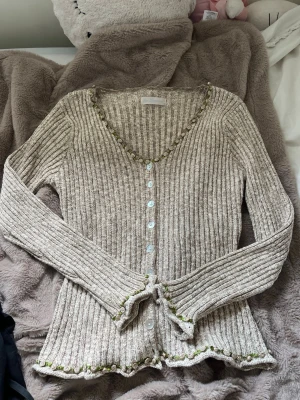 Kofta med broderade rosor - Supersöt beige ribbad kofta med v-ringning och vita knappar framtill. Koftan har grönt broderi längs kanterna och lätt utsvängda ärmar. Storlek S/M