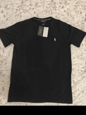 Svart t-shirt från Polo Ralph Lauren - Klassisk svart t-shirt från Polo Ralph Lauren med liten vit broderad logga på bröstet. T-shirten har rund hals och korta ärmar. Perfekt basplagg med stilren design och skön passform.   Pris kan självklart diskuteras och tröjan är helt oanvänd. Säljer på grund av att den är för liten 