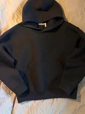 Svart hoodie från Weekday - En stilren svart hoodie från Weekday med klassisk huva och stor magficka. Hoodien har en relaxed fit och är gjord i mjukt bomullsmaterial som känns skönt mot huden. Perfekt för en chill och avslappnad look.