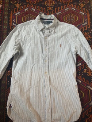 Randig skjorta från Polo Ralph Lauren - Klassisk skjorta från Polo Ralph Lauren med smala blå och vita ränder. Skjortan har lång ärm, knappar framtill och den ikoniska broderade loggan på bröstet. Tillverkad i mjuk bomull och har en tidlös, normal passform.