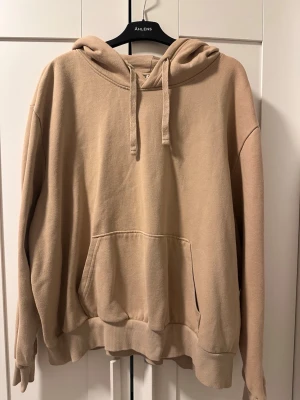 Beige oversized hoodie med ficka - Säljer en beige oversized hoodie med stor känguruficka framtill och huva med dragsko. Hoodien har ribbade muddar vid ärmslut och nederkant. Perfekt för en chill och avslappnad stil. Materialet känns mjukt och skönt mot huden.