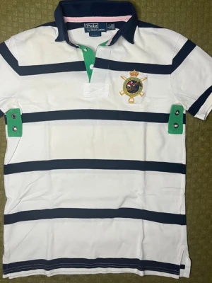 Randig pikétröja från Ralph Lauren Chief Keef - Säljer en vit och marinblå randig pikétröja från Ralph Lauren med broderad logga på bröstet. Tröjan har klassisk krage i marinblått med rosa och gröna detaljer samt knappar vid halsen. Perfekt för en sportig och stilren look.