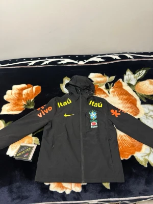 Brazil windbreaker  - Nike Brasilien Gore-Tex-jacka 🔥 Officiell landslagsjacka med högkvalitativ Gore-Tex-teknologi som håller dig torr och bekväm i alla väder. Stilren svart design med Brasilien-emblem och sponsorloggor. Ett måste för samlare, fotbollsfans eller den som vill ha en premiumjacka.