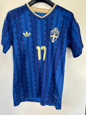 Blå Sverige fotbollströja Adidas - Säljer en blå fotbollströja från Adidas med Sveriges landslagsmärke och nummer 17, Gyökeres på ryggen. Tröjan har gul logga, detaljer och randigt mönster i olika blå nyanser. Korta ärmar och klassisk sportig passform. Perfekt för match eller träning.