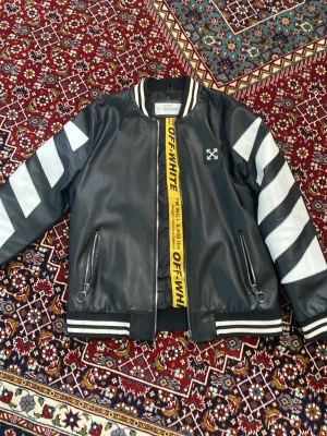 Svart bomberjacka från Off-White - Svart bomberjacka i skinnimitation från Off-White med vita ränder på ärmarna, broderad logga fram och stor vit pil-logga samt texten 'WHITE' på ryggen. Jackan har ribbade muddar och dragkedja samt en gul Off-White-rem som detalj. (Liten i storleken)