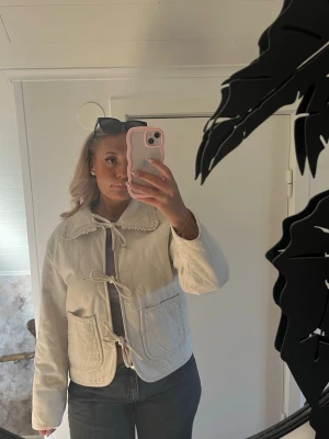 Beige vårjacka  med knytband - Säljer en beige jacka i bomull med dekorativ krage och knytband framtill. Jackan har två stora fickor, quiltad struktur och långärmade ärmar. Loose passform och stilren design som sticker ut med detaljer som snörning och broderad krage.💓