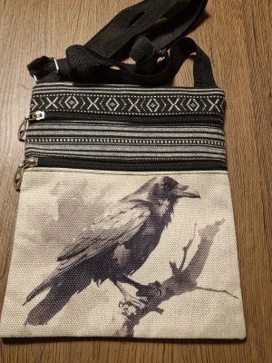 Axelväska med korpmotiv och mönster - Snygg axelväska i tyg med svartvitt korpmotiv på framsidan och etniskt inspirerat mönster upptill och på baksidan. Väskan har två dragkedjefack och justerbar axelrem. Storlek: 19 cm. Höjd 23,5 cm. Perfekt för dig som gillar unika accessoarer med naturtema.