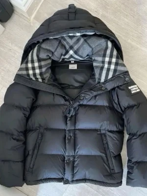 Svart dunjacka från Burberry - Snygg svart dunjacka från Burberry med klassiskt rutigt foder i huvan. Jackan har dragkedja och knappar framtill, två sidofickor och en stor huva. Perfekt för kalla vinterdagar och riktigt stilren look.