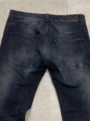 Mörkblå jeans från Nudie Jeans - Säljer ett par mörkblå jeans från Nudie Jeans med klassisk femficksmodell och subtila slitningar. Jeansen har raka ben och är tillverkade i denim med snygg tvätt och detaljerade sömmar på bakfickorna. Perfekta för en avslappnad och stilren look.