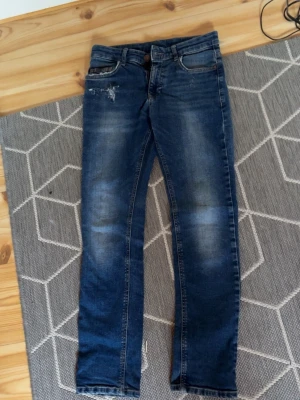 Blå slim jeans med slitningar - Ett par blå slim jeans med slitningar vid fickan för en cool, avslappnad look. Jeansen har klassisk femficksdesign och är tillverkade i denim med lätt tvättad effekt. Perfekta för dig som gillar en trendig och casual stil. pris går att diskutera. strl är i barn 158 men passar 158-168