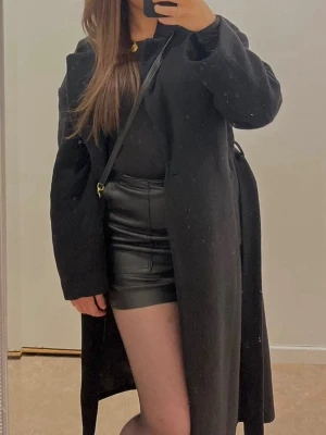 Svart kort kjol i skinnimitation - Säljer en svart kort kjol i skinnimitation med stora fickor framtill. Kjolen har en rak passform och är perfekt för dig som vill ha en edgy och trendig look. Enkel att matcha med både stickade tröjor och t-shirts.