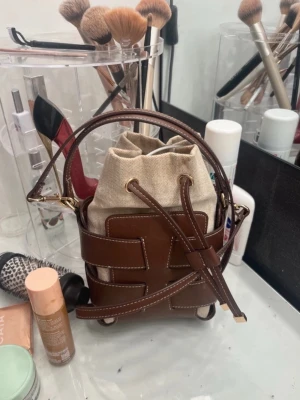 viral slutsåld väska från HM - Cool axelväska med brun läderlook och beige canvaspåse som stängs med snörning. Väskan har ett flätat mönster och justerbar axelrem med guldiga detaljer. Perfekt för dig som gillar trendiga accessoarer och vill sticka ut.