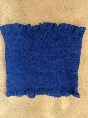 Blå bandeau tubtopp med volang - En blå bandeau tubtopp med smockad struktur och volangkant upptill och nertill. Perfekt för dig som vill ha en enkel men söt topp till sommaren. Toppens material känns stretchigt och bekvämt.