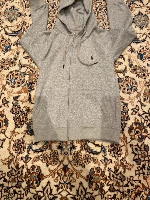 Grå zip hoodie från Polo Ralph Lauren - Snygg grå hoodie med dragkedja från Polo Ralph Lauren. Klassisk huva med dragsko och den ikoniska broderade loggan på bröstet. Tillverkad i mjukt sweatshirtmaterial som är perfekt för chill dagar. Två fickor framtill för extra stilpoäng.