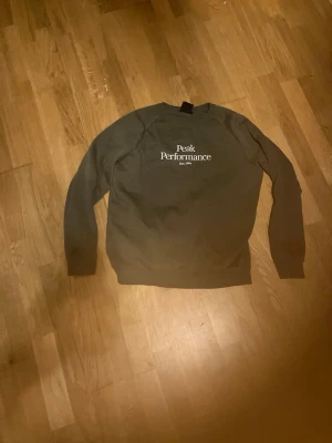 Grön sweatshirt Peak Performance - Mörkgrön sweatshirt från Peak Performance med vit logga och text på bröstet. Klassisk rund halsringning och långa ärmar. Tröjan har ribbade muddar vid ärmslut och nederkant. Perfekt för en avslappnad och stilren look.
