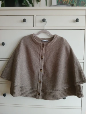 Beige poncho från Zara med knappar - Fint skick,  använd bara en.  Poncho i mjuk beige stickad ullmix från Zara. Modellen har rund halsringning, ribbstickade kanter och stora bruna knappar framtill. Perfekt för lager-på-lager och ger en chill vibe till din outfit.