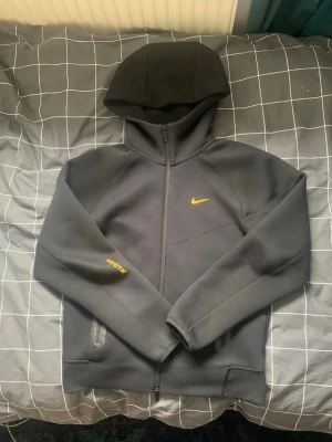 Nike x NOCTA svart hoodie med dragkedja - Svart hoodie från Nike x NOCTA med hel dragkedja, gul Nike-logga på bröstet och NOCTA-tryck på ärmen. Utrustad med två fickor med dragkedja och en stor huva. Tillverkad i ett mjukt och tjockt material som passar perfekt för en sportig och stilren look.