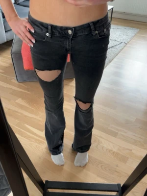 Svarta slitna jeans med hål - Säljer ett par svarta jeans med stora slitningar över knäna och lätt utsvängda ben. Jeansen har klassisk femficksmodell och låg midja. Perfekta för en avslappnad och trendig look.