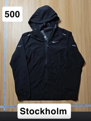 Nike Miller  - Snygg svart vindjacka från Nike med huva och dragkedja framtill. Jackan har reflekterande detaljer på axlar och ärmar samt en diskret Nike-logga på bröstet. Tillverkad i lätt och tåligt material som passar perfekt för blåsiga dagar.