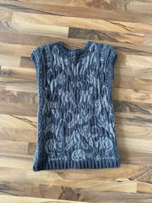 Grå stickad väst 100% ull - Snygg grå stickad väst med korta ärmar och rund hals. Västen har ett tydligt strukturmönster och ribbstickad nederkant. Perfekt att styla över en skjorta eller t-shirt för en trendig lager-på-lager-look. Kattmönster. 100% ull handstickade grå brun