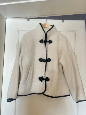 Vit teddyjacka med svarta detaljer - Snygg vit teddyjacka med svarta kantband och knäppning med tre dekorativa toggleknappar framtill. Jackan har hög krage och två fickor på sidorna. Perfekt för dig som gillar en trendig och mysig look.