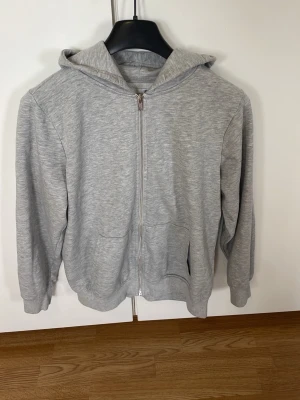 Grå zip hoodie med huva - En enkel och stilren grå hoodie med dragkedja framtill och två fickor. Hoodien har en klassisk passform, ribbade muddar vid ärmar och nederkant samt en stor huva. Perfekt för en avslappnad look. Materialet känns mjukt och bekvämt, troligen bomullsblandning.