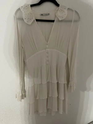Vit volangklänning från Zara - Söt vit kortklänning från Zara med volanger, v-ringning och dekorativa knappar framtill. Klänningen har långärmade ärmar med rynkade muddar och en spetsig krage med broderade detaljer. Perfekt för sommarens alla tillfällen.