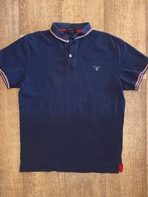 Mörkblå Piké från Gant M - Snygg marinblå pikétröja från Gant med korta ärmar och klassisk krage. Tröjan har röda och vita detaljer på krage och ärmslut samt broderad logga på bröstet. Perfekt för en stilren och sportig look. Storlek M. Snabb Leverans!