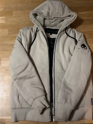 Moose knuckles Bunny - Snygg beige vindjacka med huva från Moose Knuckles. Jackan har svart dragkedja, ribbade muddar och mjukt foder i pälsimitation. Detaljerad med logotyp på ärmen och svarta kontrastdetaljer vid dragkedjorna. Perfekt för kyliga dagar. ÄKTA jacka