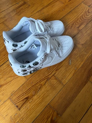 Vita sneakers med svart mönster - Säljer ett par vita sneakers med svart grafiskt mönster på hälen och sidan. Skorna har klassisk låg profil, perforerad tå och vita skosnören. Tillverkade i syntetmaterial med gummisula. Perfekta för dig som vill sticka ut med en unik detalj.