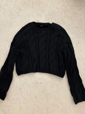 Svart kabelstickad cropped tröja - Svart kabelstickad tröja från Bik Bok med croppad passform och rund halsringning. Tröjan har långa ärmar och ribbade muddar vid ärmslut och nederkant. Perfekt för lager-på-lager och enkel att styla till olika outfits. (Lite öppen rygg)