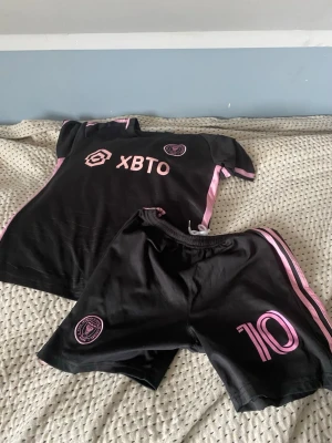Inter Miami svart fotbollskit rosa detaljer från Adidas - Säljer ett svart fotbollskit från Inter Miami CF med rosa detaljer. Tröjan har klubbmärke och sponsorlogga framtill, Adidas, och shortsen har nummer 10 samt klubbmärke. Båda delarna har rosa ränder längs sidorna. Perfekt för träning eller match. 89 kr för båda, 75 kr st annars. Passar för 10-12 år. 